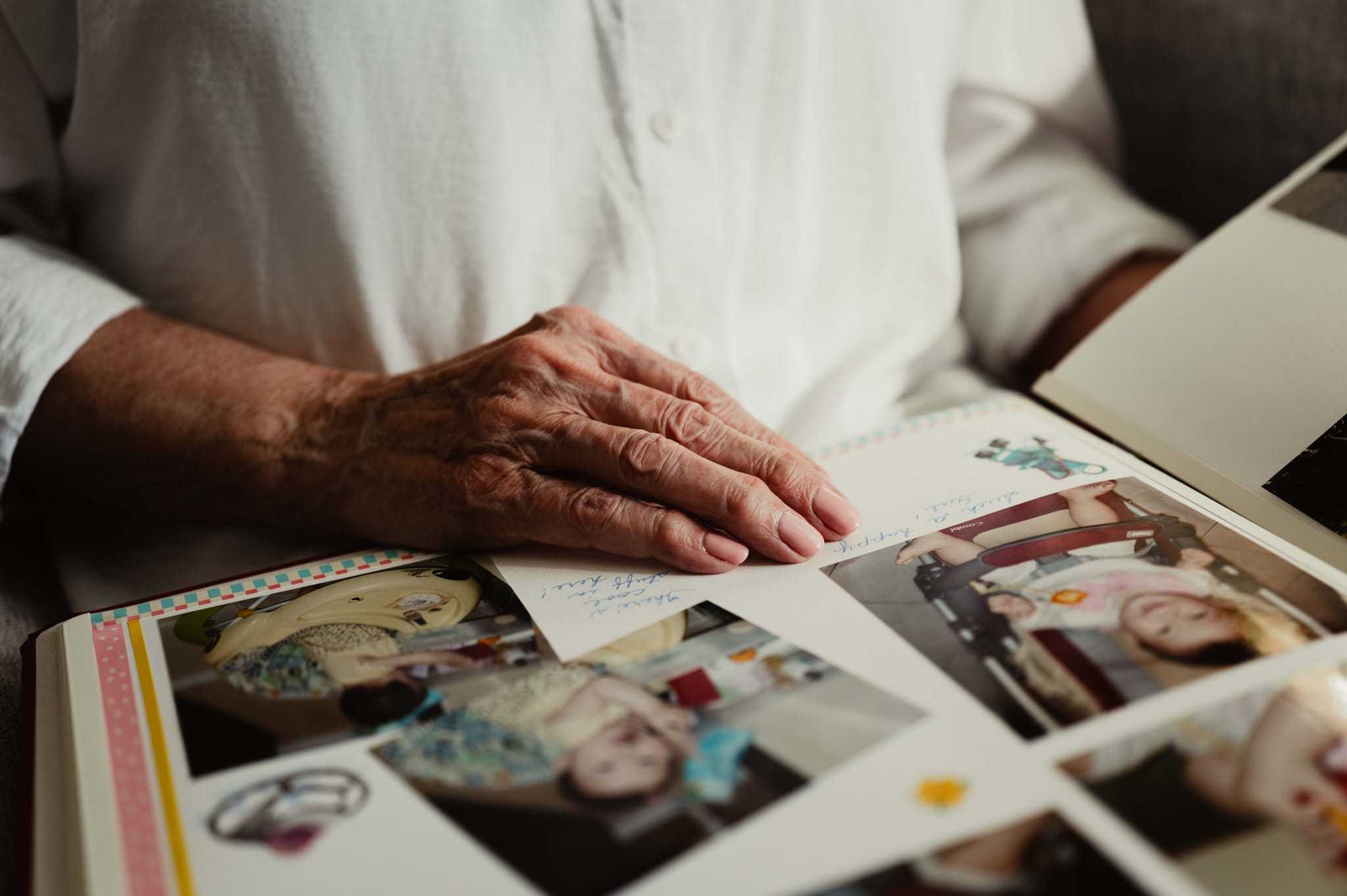 An elderly woman's hand resting on an open family photo album filled with old snapshots — honouring a lifetime of stories in Lethbridge
